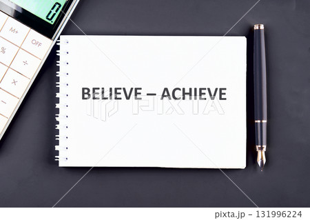 A Notepad with the words BELIEVE - ACHIEVE rests on a dark surface, next to a pen and a calculator, embodying goals, planning and success at Work 131996224