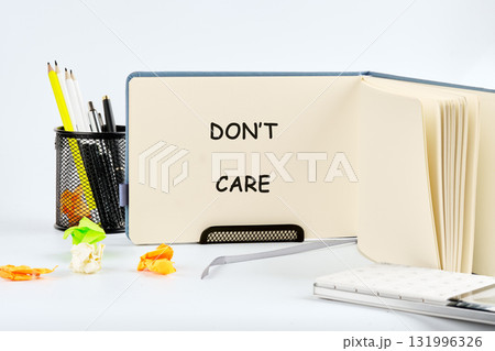The scene contains an open notebook displaying the words Dont Care. Nearby are writing utensils in a metal container, plus crumpled paper, and a notebook, all on a white surface The scene contains an open notebook displaying the words Dont Care. Nearby are writing utensils in a metal container, plus crumpled paper, and a notebook, all on a white surface 131996326
