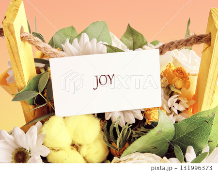 Assortment of yellow and white flowers arranged in a decorative wooden basket with greenery and a message card saying Joy 131996373