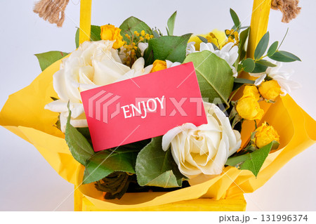 A yellow basket brims with white roses and yellow daisies. A coral card reads ENJOY, suggesting a thoughtful gift for someone special, promoting happiness and appreciation 131996374