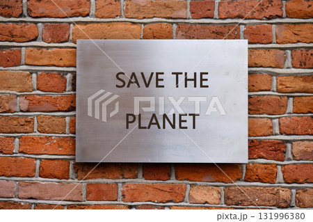 A metal sign with the words SAVE THE PLANET is affixed to a rustic brick wall. Sunlight casts shadows, emphasizing the urgent message of environmental stewardship 131996380