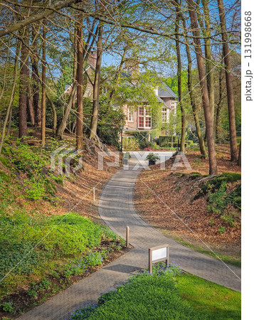 A winding paved path slopes down through the forest leading towards a rustic house nestled in the woods. Peaceful scene with secluded atmosphere A winding paved path slopes down through the forest leading towards a rustic house nestled in the woods. Peaceful scene with secluded atmosphere 131998668