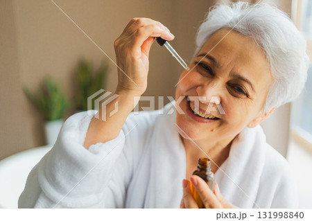 Woman applying natural drops cosmetics oil to face skin. Mature adult old woman applying nourishing anti age serum with pipette doing daily beauty routine. Senior woman gets kin scare facial treatment 131998840