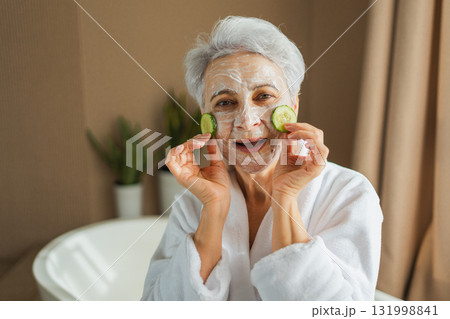 Mature adult old woman with nourishing anti age face cream mask holding cucumber slices. Senior woman with white mask creme on skin gets facial treatment daily beauty routine. Skin care aging beauty Mature adult old woman with nourishing anti age face cream mask holding cucumber slices. Senior woman with white mask creme on skin gets facial treatment daily beauty routine. Skin care aging beauty 131998841