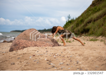A determined woman is seen energetically pushing a large, heavy rock on a sandy beach with a beautiful, scenic backdrop 132001547