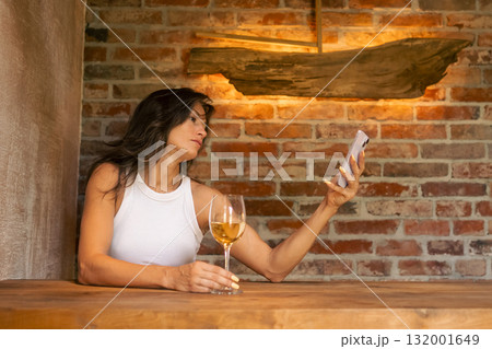 In a comfortable and cozy rustic environment, a woman enjoys relaxation while sipping her wine and using her smartphone In a comfortable and cozy rustic environment, a woman enjoys relaxation while sipping her wine and using her smartphone 132001649