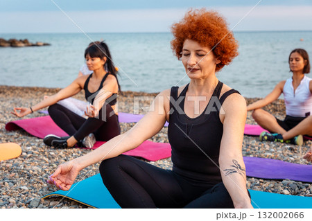 Portrait of adult Caucasian red haired woman meditate sitting on sports mat on pebble beach. Concept of outdoor yoga class 132002066