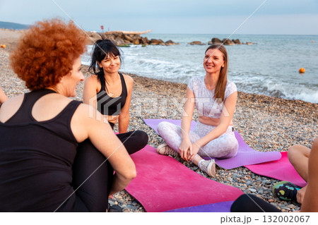 Group of smiling young-adult multi-racial women are sitting on sports mats on wild beach and talking to each other. Concept of female circle of communication and support class 132002067