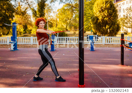 Training in park. Side view of adult Caucasian woman looking in camera does stretching with elastic band on sports playground. Healthy lifestyle 132002143