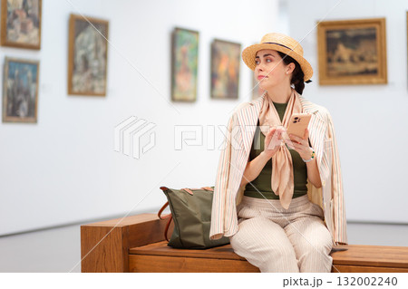 Mid shot of young Caucasian beautiful woman in an elegant suit is sitting on a bench in an art gallery and using her smartphone. Copy space. Modern cultural education Mid shot of young Caucasian beautiful woman in an elegant suit is sitting on a bench in an art gallery and using her smartphone. Copy space. Modern cultural education 132002240