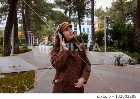 Young beautiful Caucasian woman in a coat and beret walks through the autumn city listening to music in headphones and enjoying a moment of relaxation during her day off. 132002299