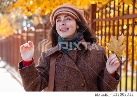 Portrait of young pretty Caucasian happy woman with braces on her teeth in a coat and hat holds bunch of autumn yellow leaves and waving hands, standing on the street. Lifestyle and urban life Portrait of young pretty Caucasian happy woman with braces on her teeth in a coat and hat holds bunch of autumn yellow leaves and waving hands, standing on the street. Lifestyle and urban life 132002313