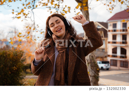 Mid shot of beautiful happy young Caucasian woman in a coat, smiling and listening to music with headphones and dancing in an autumn park. Concept of recreation and psychology Mid shot of beautiful happy young Caucasian woman in a coat, smiling and listening to music with headphones and dancing in an autumn park. Concept of recreation and psychology 132002330