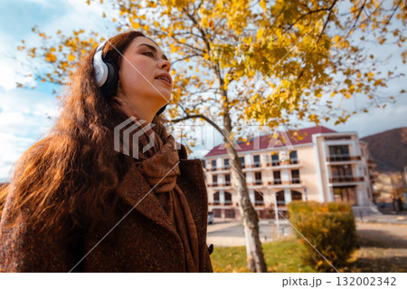 Bottom view of a Caucasian young woman in a coat listening to music with headphones in autumn park. Concept of happiness and carefree Bottom view of a Caucasian young woman in a coat listening to music with headphones in autumn park. Concept of happiness and carefree 132002342