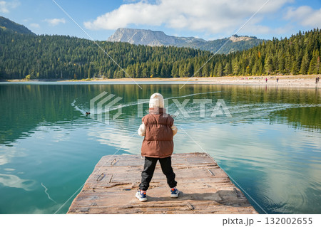 Vertical photo of boy standing on wooden pier and looking at Black Lake surrounded by pine forest and mountains in Durmitor National Park, Montenegro. Tranquil natural landscape with reflections on 132002655