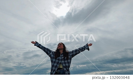 A beautiful woman with long dark hair is facing the camera with the sky in the background, raising her hands to the sky and enjoying the nature and the moment. 132003048