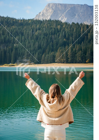 Half-length view of a woman standing on a wooden pier with raised arms, enjoying the scenic Black Lake and forest landscape in Durmitor National Park, Montenegro. 132003138