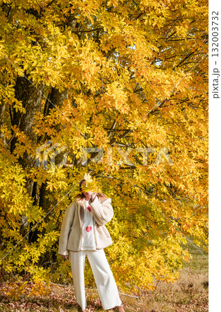 Scenic view of a woman standing under a yellow autumn tree, hiding her face behind fallen leaves. Warm fall colors create a peaceful seasonal atmosphere. 132003732