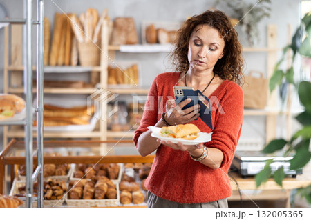 European female customer with phone takes pictures of burger croissant in disposable plate dish European female customer with phone takes pictures of burger croissant in disposable plate dish 132005365