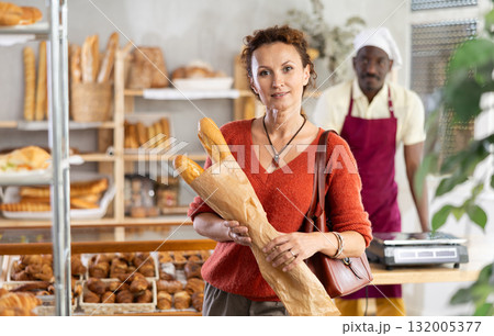 Woman buyer with pastry and baguettes in paper package stands near glass showcase in bakery 132005377