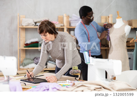 Woman tailor creates sketch of dress on tablet, man companion work in background with mannequin Woman tailor creates sketch of dress on tablet, man companion work in background with mannequin 132005599