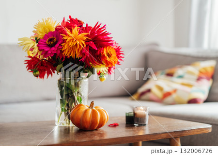 Autumn, fall cozy mood composition for hygge home decor. Bouquet of fresh multicolor dahlia flowers, small pumpkin, burning candles on the coffee table in modern living room. Selective focus. 132006726