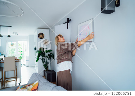 Side view middle aged woman putting painting picture in frame on the wall. Casual lady taking care of coziness in her new stylish apartment. Mature female decorating room. Moving and renovate house Side view middle aged woman putting painting picture in frame on the wall. Casual lady taking care of coziness in her new stylish apartment. Mature female decorating room. Moving and renovate house 132006751