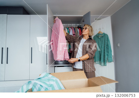Middle aged woman selecting clothes from wardrobe for donating to Charity shop. Decluttering, Sorting clothes and Cleaning Up. Reuse, second-hand concept. Conscious consumer, sustainable lifestyle. 132006753
