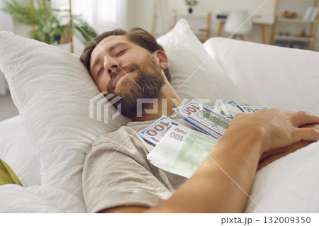 Smiling young man sleeping on bed with a bunch of cash dollars keeping his money savings at home. Smiling young man sleeping on bed with a bunch of cash dollars keeping his money savings at home. 132009350