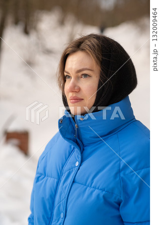 A woman wearing a blue winter coat and black headscarf looks thoughtfully into the distance against a snowy backdrop filled with trees. 132009464