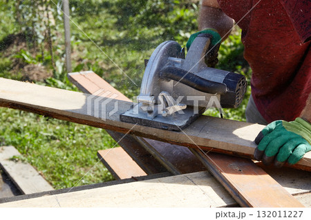 Skilled worker using a circular saw to cut wooden planks in a sunny outdoor setting Skilled worker using a circular saw to cut wooden planks in a sunny outdoor setting 132011227