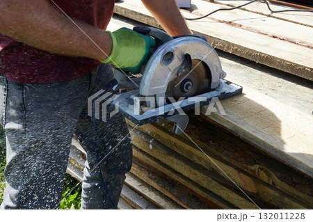 A worker saws wooden planks with a circular saw in a green, sunny place. A worker saws wooden planks with a circular saw in a green, sunny place. 132011228