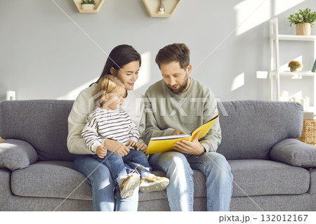 Happy family sitting on sofa reading a book together in their living room Happy family sitting on sofa reading a book together in their living room 132012167