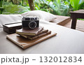 Vintage camera and leather-bound journals resting on a daybed in a lush, tropical setting. Perfect moment for travel, reflection, and creative escape 132012834