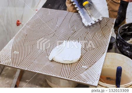 Worker spreads adhesive onto large tile, preparing it for installation on floor. Worker spreads adhesive onto large tile, preparing it for installation on floor. 132013309