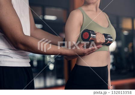 Young asian woman doing exercises with dumbbells in a gym while her trainer is supporting her. 132013358