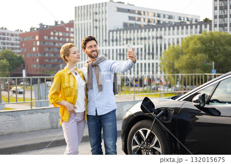 A smiling couple takes a selfie while their electric vehicle charges in an urban setting, highlighting modern eco-friendly transportation. 132015675