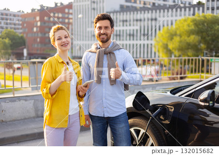 Happy couple giving thumbs up while their electric car is charging at a charging station. 132015682