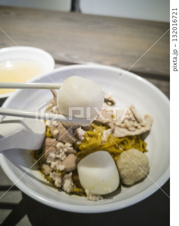 A fishball is picked up by a pair of chopsticks in the foreground against the background of a bowl of minced meat noodle A fishball is picked up by a pair of chopsticks in the foreground against the background of a bowl of minced meat noodle 132016721