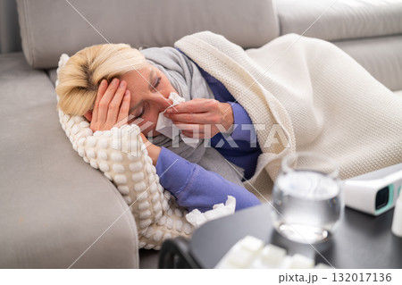 Blonde woman lying on couch with runny nose, covered with blanket, looking tired. Pills, thermometer, and tissues in the foreground. Concept of flu, illness, and home recovery. 132017136