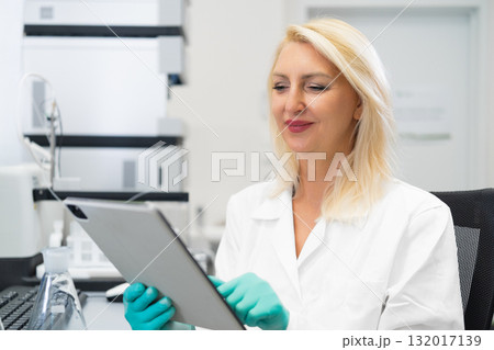 Blonde female scientist without mask and wearing rubber gloves recording experiment results on a tablet in a chemistry laboratory filled with flasks, beakers, and HPLC instruments 132017139