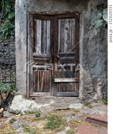 Weathered wooden door with padlock and chain on a textured concrete wall. Rustic entrance detail with green foliage. 132020331