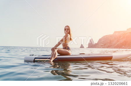 Woman Paddleboarding Ocean Coastline - Young woman in black bikini relaxing on paddleboard in ocean with coastal cliffs in background. Woman Paddleboarding Ocean Coastline - Young woman in black bikini relaxing on paddleboard in ocean with coastal cliffs in background. 132020661