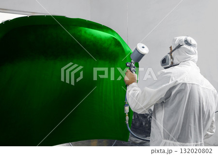 Automobile repairman painter in mask and protective workwear painting car hood to green color in paint chamber. 132020802