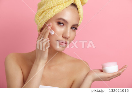 Seductive female model applies cream on cheek at pink wall 132021016