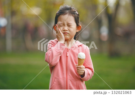 Unhappy Child Girl Touching her Cheek Feeling Toothache, Holding Ice Cream, Suffers from Dental Decay Against Summer Park Background 132021063