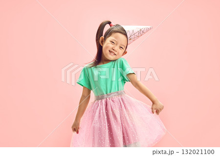 Cheerful Birthday Asian Child Girl in Festive Clothes, Having Party Hat on Head, Cute Raising Shoulders, Funny Straightens her full Skirt to the Sides Standing Over Pink Isolated Backgound. 132021100