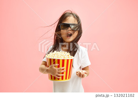 Surprised Asian Little Girl Watching Cartoon Movie in 3D Glasses and Eating Large Portion of Popcorn Standing on Pink Isolated Background. Surprised Asian Little Girl Watching Cartoon Movie in 3D Glasses and Eating Large Portion of Popcorn Standing on Pink Isolated Background. 132021102