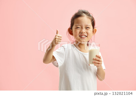 Happy Smiling Little Brunette Child Girl Drinking Milk From Glass With a Trace Frm Milk on Lips, Expresses Pleasure Positively Showing Thumb up Standing On Pink Isolated 132021104