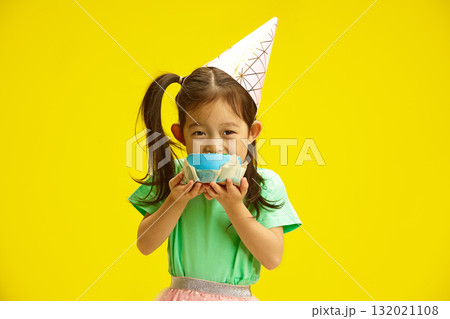 Cheerful Child girl in Party Hat Holding and Bites Piece From Circle Birthday Cake Standing Against Yellow Background. 132021108